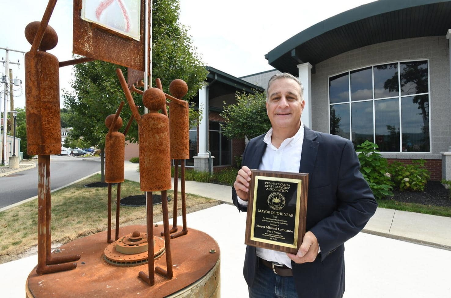 Pittston Mayor Mike Lombardo smiles and holds a plaque outside next to a metal sculpture